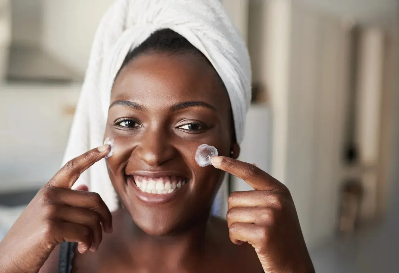 Donna sorridente con turbante applica crema sul viso, un'immagine che evoca il desiderio di vedere i risultati del **retinolo prima e dopo**.