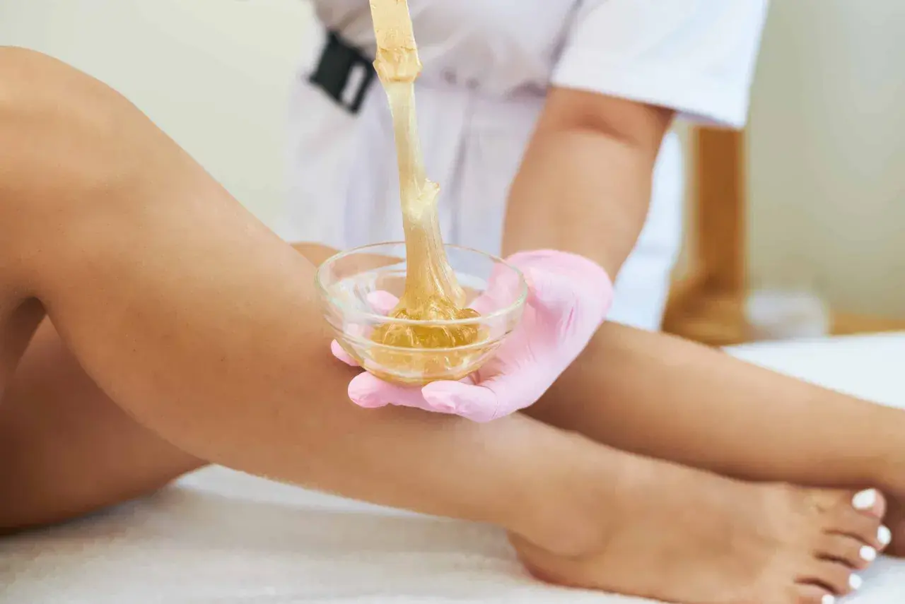 Una mano con guanto rosa preleva cera da una ciotola per la depilazione femminile delle gambe.