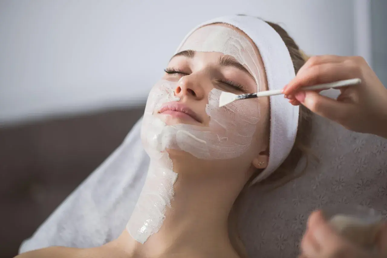 Una donna riceve un peeling viso. La maschera bianca viene applicata con un pennello, mostrando come si fa un trattamento di bellezza.