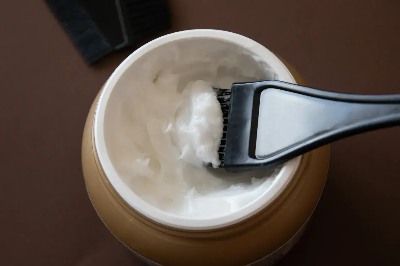 Un pennello raccoglie una crema bianca da un barattolo dorato, ideale per la cura dei capelli con burro di karitè.