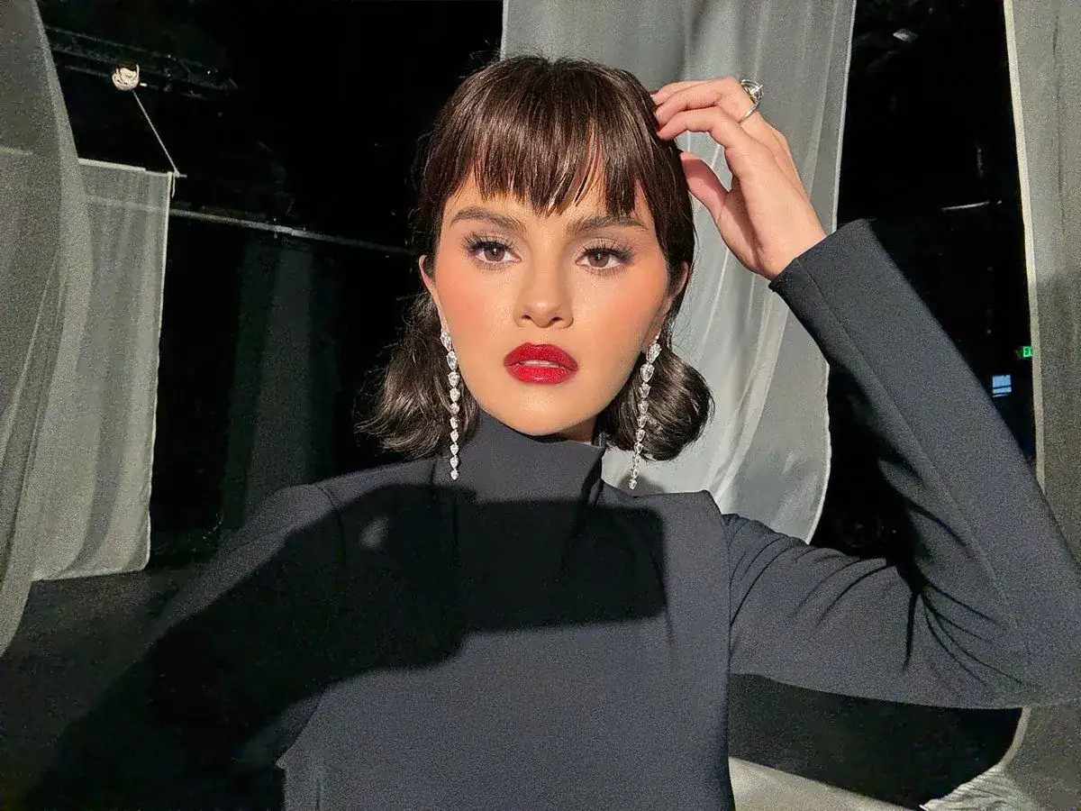 Donna con un elegante abito nero e una frangia corta che incornicia il viso, illuminato da un rossetto rosso acceso e orecchini pendenti scintillanti.