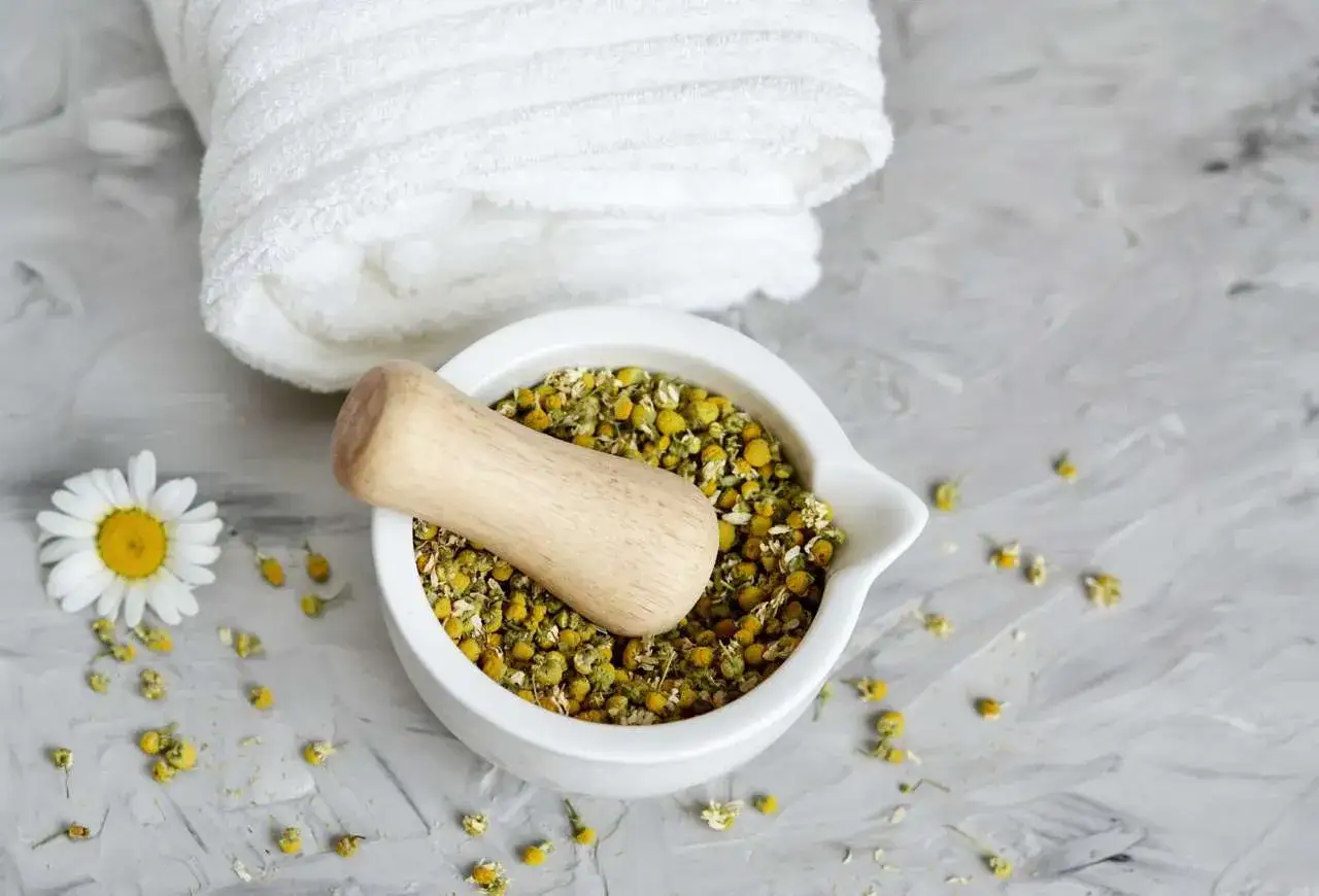 Mortaiolo con camomilla secca, un fiore fresco e un asciugamano bianco. Scopri gli shampoo alla camomilla effetti benefici.