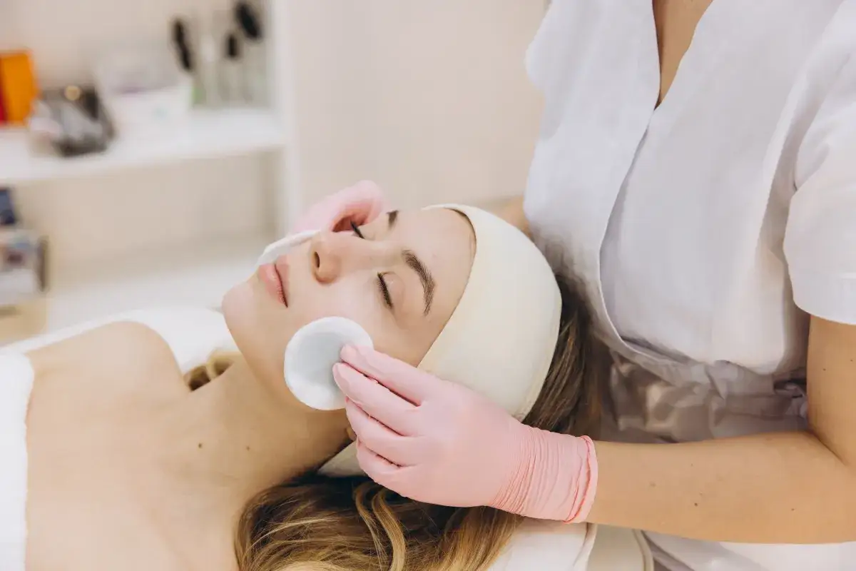 Una donna riceve un trattamento di pulizia del viso con dischetti di cotone. Le mani guantate rosa accarezzano delicatamente la pelle.