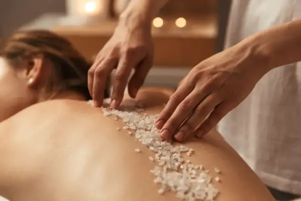 Mani esperte applicano un scrub corpo con cristalli di sale sulla schiena di una persona, un rituale di benessere prima della doccia.