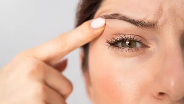 Dettaglio di un occhio incappucciato con trucco, evidenziato da un dito che solleva la palpebra.