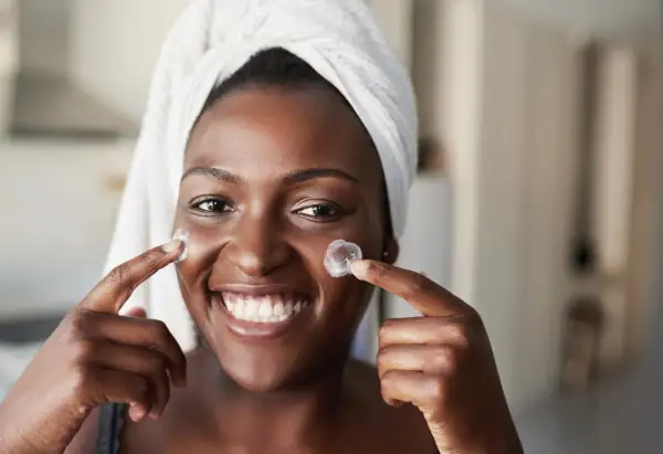 Donna sorridente con turbante applica crema sul viso, un'immagine che evoca il desiderio di vedere i risultati del **retinolo prima e dopo**.