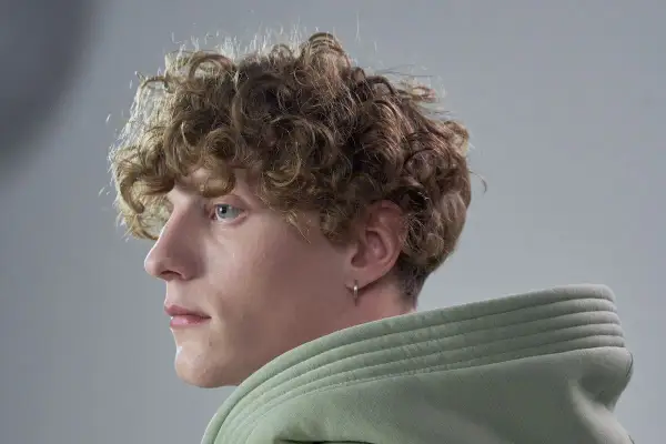 Primo piano di un giovane con capelli mossi uomo, occhi azzurri e un orecchino a cerchio. Indossa una felpa verde chiaro.