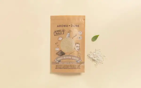 Confezione di maschera argilla bianca Aroma-Zone, con polvere sparsa e fogliolina verde su sfondo chiaro.